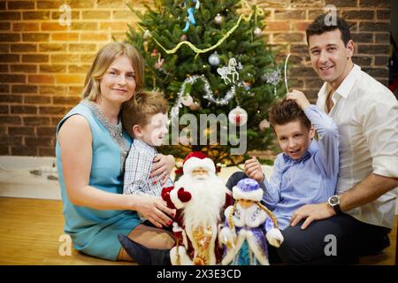 Una famiglia felice di quattro persone siede sul piano in camera sotto l'albero di natale con giocattoli come padre Frost e Fanciulla delle nevi. Foto Stock