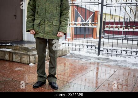 L'uomo in uniforme militare si trova vicino all'ufficio del checkpoint e alla recinzione metallica il giorno d'inverno. Foto Stock