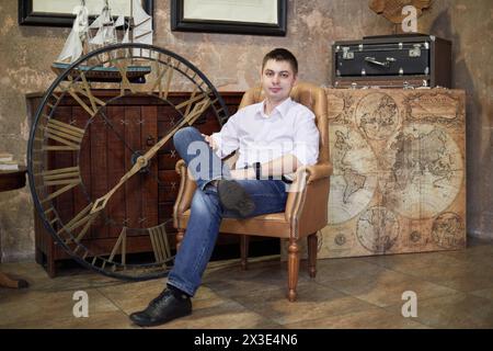 Un giovane uomo siede in poltrona in camera con una vecchia mappa, una nave simulata, valigie da viaggio. Foto Stock