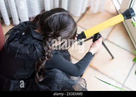 Ragazza con lunghe trecce in un abito nero guarda attraverso un telescopio Foto Stock