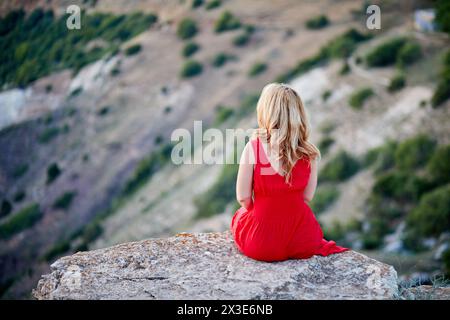 Donna in abito rosso seduta sul bordo di una scogliera di pietra, vista posteriore. Foto Stock