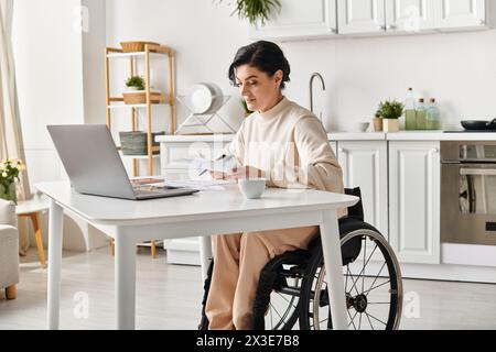 Una donna disabile su una sedia a rotelle che lavora in remoto su un notebook dalla sua cucina. Foto Stock