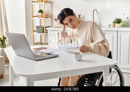 Una donna disabile su una sedia a rotelle lavora con il suo laptop a un tavolo nella sua cucina. Foto Stock