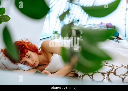 giovane donna dai capelli rossi giace nel letto sotto la coperta, usa lo smartphone, concetto di vacanza mattutina Foto Stock