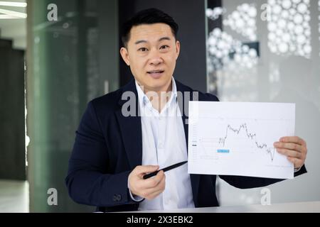 Un uomo d'affari professionista dell'Asia orientale che presenta con fiducia un grafico finanziario, interagendo direttamente con la fotocamera in un ambiente d'ufficio contemporaneo. Foto Stock