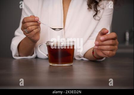 Le mani di una donna in primo piano che tiene un cucchiaino con zucchero bianco sopra una tazza di tè nero appena fatto nella cucina di casa, preparando una bevanda calda per la colazione Foto Stock