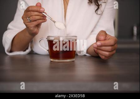 Primo piano delle mani della donna con un cucchiaino di zucchero, dolcificando la bevanda in un bicchiere. Le mani di una donna tengono un cucchiaio con zucchero. Aggiunta di zucchero a Foto Stock