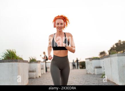 Donna che ascolta la musica con le cuffie mentre fa jogging sul sentiero. Jogger Runner dalla vestibilità sorridente in abbigliamento sportivo che si allenano all'aperto. Giovane donna attiva che corre sotto il cielo limpido. Stile di vita sano. Foto Stock