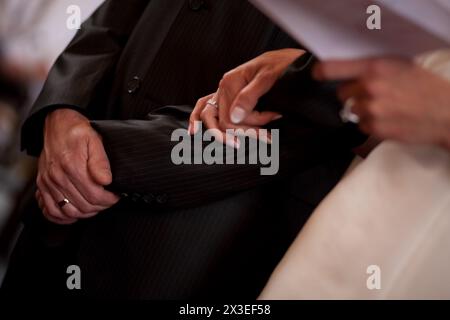 Questo primo piano cattura la sottile intimità di una coppia durante le promesse nuziali. La mano dello sposo, adornata da una banda nuziale, regge delicatamente la sposa, mostrando il suo anello di fidanzamento. L'attenzione alle mani sullo sfondo formale dell'abbigliamento simboleggia la tenerezza e la promessa del momento. Momento intimo: Primo piano della presa di mano di una coppia amorevole durante le promesse. Foto di alta qualità Foto Stock