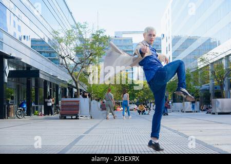 giovane gay con i capelli biondi corti, di profilo saltando in strada alzando la gamba e sventolando la giacca con le mani, guardando la macchina fotografica, le vite Foto Stock