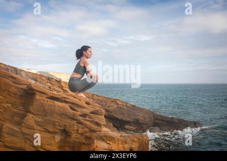 Donna sportiva seduta su una roccia che guarda al mare, rilassandosi dopo l'allenamento o lo yoga Foto Stock