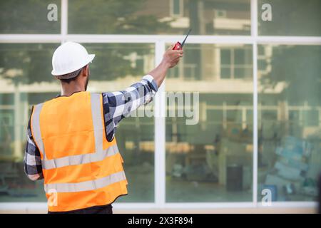 Il caposquadra dietro le quinte che indossa giubbotto riflettente e elmetto ispeziona il cantiere per assicurarsi che la costruzione soddisfi i piani di progettazione del team di progettazione Foto Stock