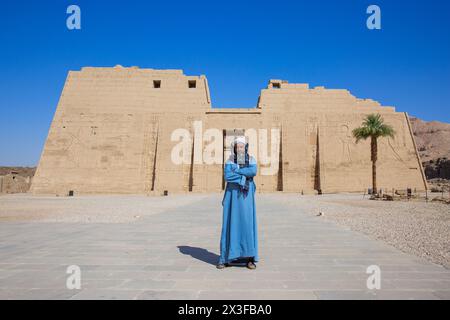 Un uomo egiziano in abbigliamento tradizionale al primo pilone del tempio mortuario di Ramses III a Medinet Habu sulla sponda occidentale di Luxor, in Egitto Foto Stock