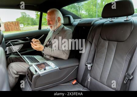 Un signore anziano in tuta esamina i messaggi sul suo cellulare durante un viaggio in auto, il suo portatile nelle vicinanze Foto Stock