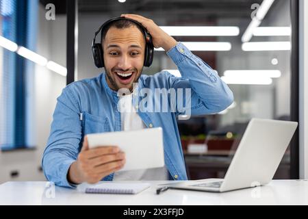 Felice e sorpreso giovane musulmano seduto in ufficio al tavolo con le cuffie e guardando entusiasticamente lo schermo del tablet, tenendo la testa con la mano. Foto Stock