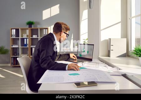 Ispettore catastale seduto alla scrivania dell'ufficio, lavorando con le mappe catastali e utilizzando un computer portatile Foto Stock