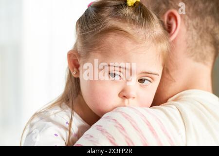 Primo piano di papà che tiene in mano una piccola figlia carina con la sindrome di Down che guarda lontano dalla telecamera, copia spazio Foto Stock