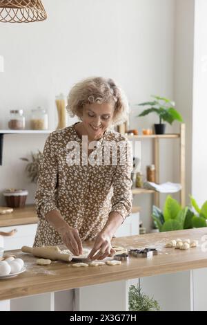 Una bella donna sorridente di mezza età prepara biscotti con impasti fatti in casa Foto Stock