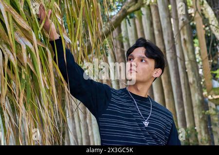 Un uomo con una camicia a righe bianca e nera e una collana con catena d'argento è in piedi. Il ragazzo è con la mano sollevata toccando i rami di un albero. Foto Stock