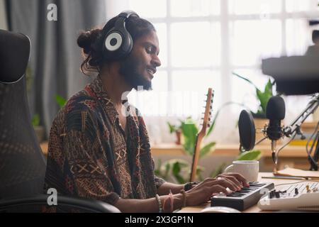 Felice giovane cantante e cantautore premendo i tasti del pianoforte elettrico e eseguendo una nuova canzone mentre la registrava dal posto di lavoro in studio Foto Stock