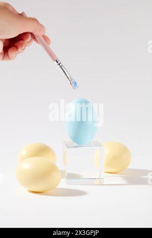 Un uovo di Pasqua di colore blu è posto su un podio di vetro a forma di cubo, circondato da altre uova gialle. Una mano che regge un pennello per dipingere l'uovo su di te Foto Stock