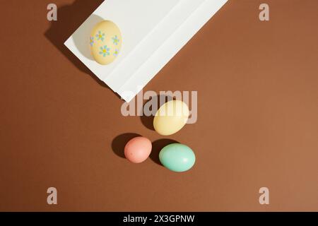 Vista dall'alto di diverse uova di Pasqua di diverse dimensioni e colori sono decorate su sfondo marrone. Un uovo posto su una scala bianca. Giorno di Pasqua a C Foto Stock