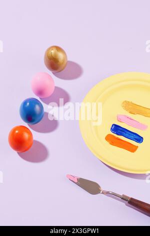 Una vivace collezione di uova di Pasqua colorate disposte in fila, esposte con un piatto giallo contenente vari colori di vernice. Una tavolozza di coltelli decorativi Foto Stock