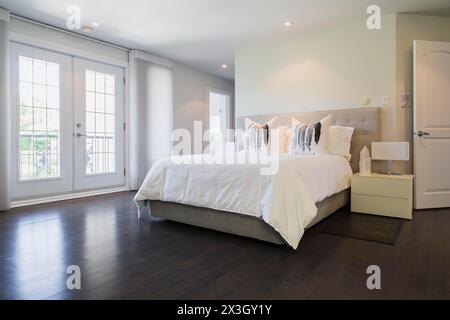 Camera da letto principale con letto queen size, comodini laccati bianchi e pavimento in legno scuro al piano superiore all'interno di una lussuosa casa residenziale Foto Stock