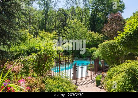 In terreno piscina circondata da color rame ferro battuto metallo recinzione di sicurezza e varie piante perenni, arbusti e alberi nel cortile del Foto Stock