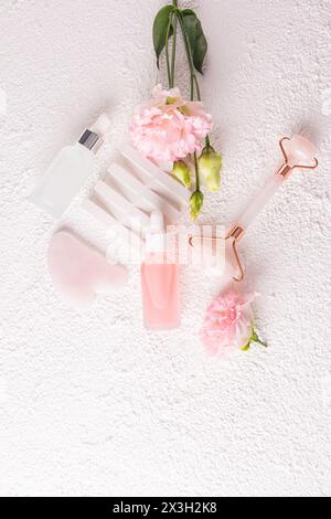 Set di cosmetici per massaggi e cura della pelle di viso e collo su sfondo bianco con fiori rosa. Due bottiglie di siero e olio. Massaggiatore. gua sha Foto Stock