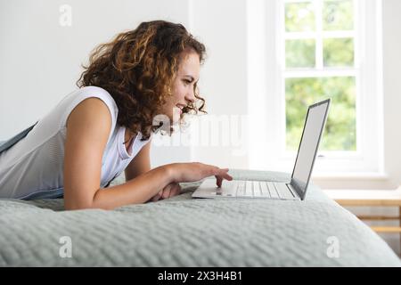Giovane donna caucasica sdraiata a letto a casa, usando un computer portatile Foto Stock