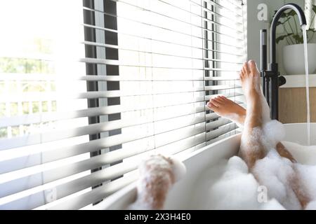 Una giovane donna caucasica che si rilassa nella vasca da bagno di casa, che sbircia i piedi attraverso le tapparelle, copia spazio Foto Stock