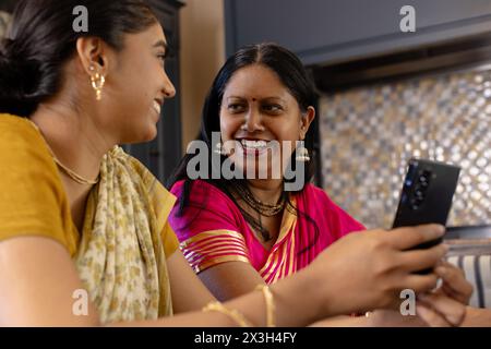 Madre e figlia indiane, sorridono a casa, guardano lo smartphone Foto Stock