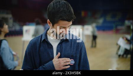 Felice elettore maschile, cittadino americano dopo aver votato durante le elezioni presidenziali negli Stati Uniti. Un uomo caucasico con il distintivo sorride e guarda la macchina fotografica in piedi al polling station. Dovere civico. Verticale. Foto Stock