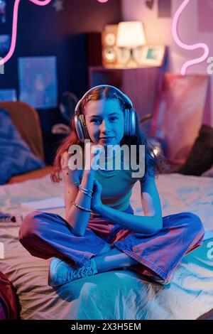 Immagine verticale di un'elegante ragazza adolescente che indossa le cuffie seduto sul letto nella sua camera da letto decorata con luce al neon a tarda sera Foto Stock