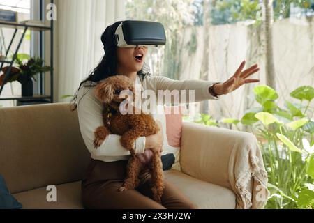 Giovane donna eccitata che tiene in mano il cane quando gioca a videogame con occhiali vr Foto Stock