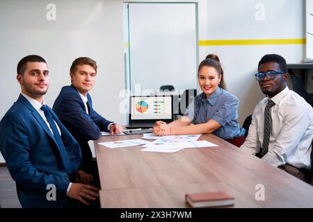 Il team multietnico si siede al tavolo in un ufficio moderno. Due uomini, due donne analizzano i dati, discutono del progetto. Membri caucasici, asiatici, africani, arabi Foto Stock