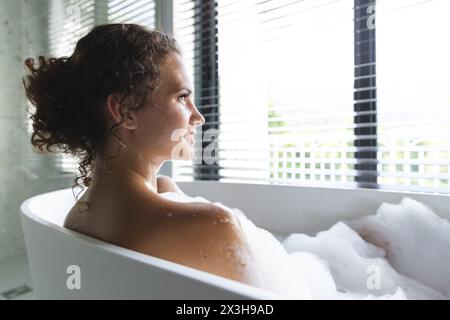 Una giovane donna caucasica che si rilassa nella vasca da bagno piena di bollicine a casa, spazio copia Foto Stock