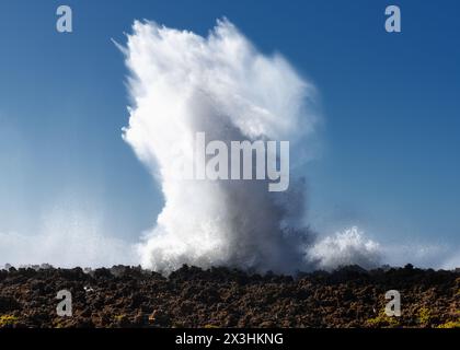 Enorme onda che si schianta su una barriera rocciosa sotto un cielo blu e crea un'eruzione d'acqua Foto Stock