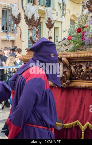 Tarragona, Spagna - 27 aprile 2024: Penitente in capirote durante la processione della settimana Santa con gradino floreale sullo sfondo, cattura spiritualità e trad Foto Stock