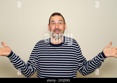 Uomo ispanico con la barba a 40 anni che indossa un maglione a righe privo di senso e confuso con le braccia aperte, nessun concetto di idea. Isolato in uno studio beige Foto Stock