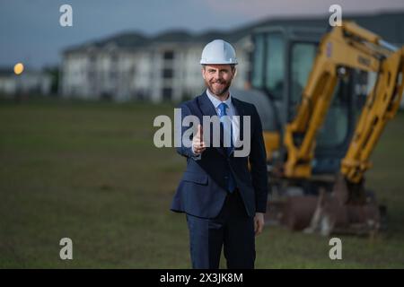 Proprietario di un'impresa edile. Uomo con tuta e cappello duro al cantiere edile. Ingegnere civile di fronte alla casa vicino all'escavatore. Arco Foto Stock