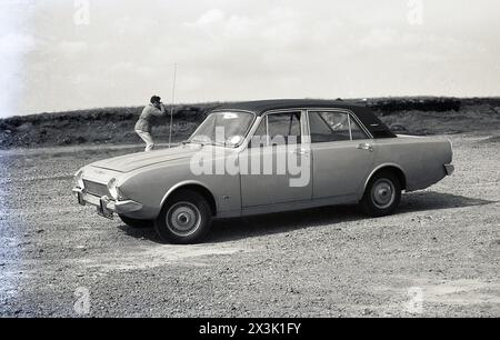 1970, storica, un'auto Ford Corsair parcheggiata fuori su terreni accidentati, il proprietario maschio fuori a scattare fotografie. La vettura fu prodotta dal 1964 - originariamente conosciuta come Ford Consul Corsair, in seguito semplicemente Ford Corsair - fino al 1970, con diverse varianti. Il modello qui è una Corsair V4 2000E, una Corsair con tetto in vinile, progettata per competere con altre auto executive più esclusive come la Rover 2000 e la Triumph 2000. Foto Stock