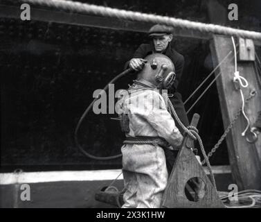 1942, storico, in tempo di guerra e uomo che indossa un berretto piatto che aiuta un subacqueo in acque profonde con l'equipaggiamento da immersione dell'epoca, in procinto di entrare in acqua, probabilmente Southampton Docks, Inghilterra, Regno Unito. Noto come abito da immersione standard, equipaggiamento da mare profondo o pesante, questo tipo di tuta da immersione è stata utilizzata per lavori subacquei profondi e consisteva in un casco in metallo pesante, rame, ottone o bronzo montato su una tuta impermeabile in tela, un tubo flessibile per aria o respirazione collegato alla superficie e pesi in piombo per contrastare il galleggiamento. Foto Stock