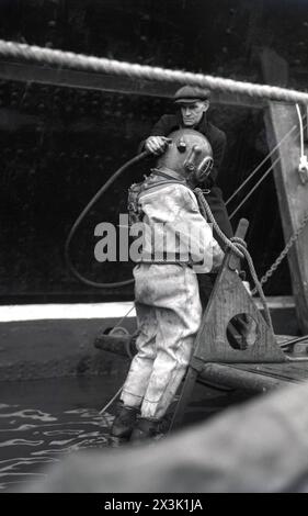 1942, storico, in tempo di guerra e uomo che indossa un berretto piatto che aiuta un subacqueo in acque profonde con l'equipaggiamento da immersione dell'epoca, in procinto di entrare in acqua, probabilmente Southampton Docks, Inghilterra, Regno Unito. Noto come abito da immersione standard, equipaggiamento da mare profondo o pesante, questo tipo di tuta da immersione è stata utilizzata per lavori subacquei profondi e consisteva in un casco in metallo pesante, rame, ottone o bronzo montato su una tuta impermeabile in tela, un tubo flessibile per aria o respirazione collegato alla superficie e pesi in piombo per contrastare il galleggiamento. Foto Stock