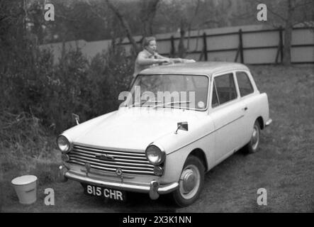 Anni '1960, storica, fuori su un prato posteriore, una donna che pulisce una piccola auto dell'epoca, una Austin A40 Farina. Prodotta dalla Britsh Motor Corporation (BMC), l'A40 fu introdotta nel 1958, con lo stile di una società italiana, Pininfarina. La vettura era in produzione, con diversi modelli, fino al 1967. Foto Stock