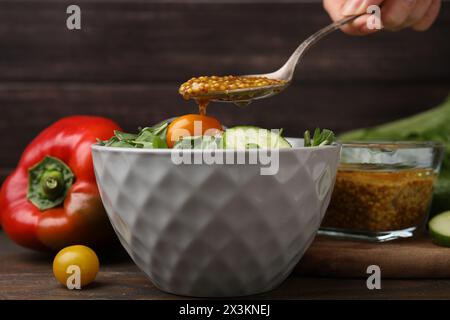 Donna che versa una gustosa salsa a base di aceto (Vinaigrette) dal cucchiaio alla ciotola con insalata al tavolo di legno, primo piano Foto Stock