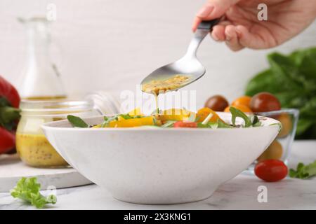 Donna che versa una gustosa salsa a base di aceto (Vinaigrette) dal cucchiaio alla ciotola con insalata sul tavolo di marmo bianco, primo piano Foto Stock