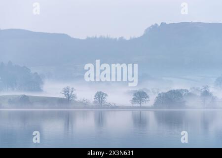Splendida immagine di paesaggio tranquillo della mattina nebbiosa di primavera su Windermere nel Lake District con barche ormeggiate sul lago e lontane cime nebbiose Foto Stock