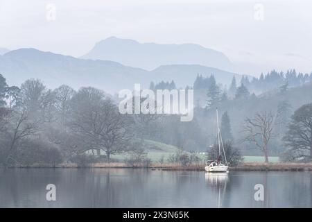 Splendida immagine di paesaggio tranquillo della mattina nebbiosa di primavera su Windermere nel Lake District con barche ormeggiate sul lago e lontane cime nebbiose Foto Stock